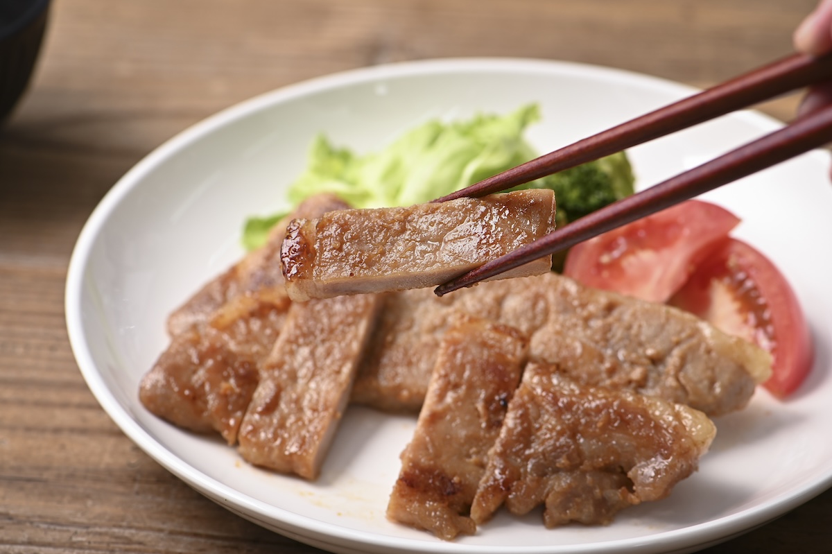 シチュエーションの撮影実績：焼いた豚肉を白い皿に盛り、赤身と脂のバランスがよい一切れを箸で持ち上げて撮影しました。